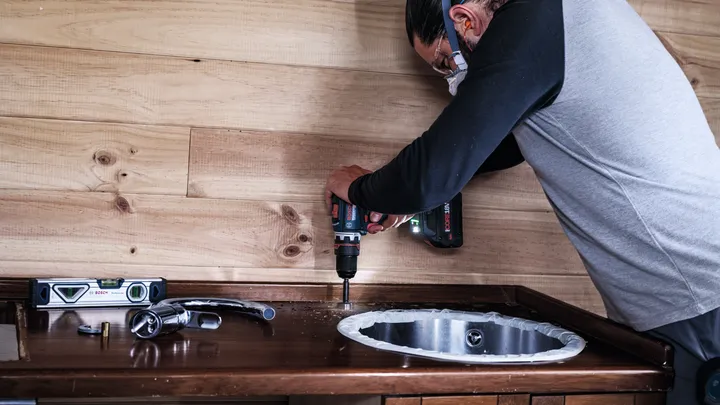 Person wearing safety equipment drills into wood beside a sink in a kitchen workspace.
