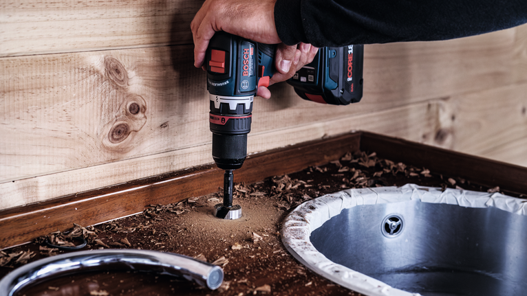 Person using a power drill to create a hole in a wooden countertop near a sink.