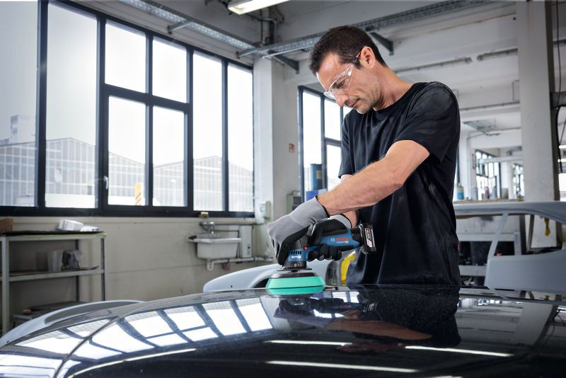 A person wearing safety equipment polishes a car hood with a cordless polisher.