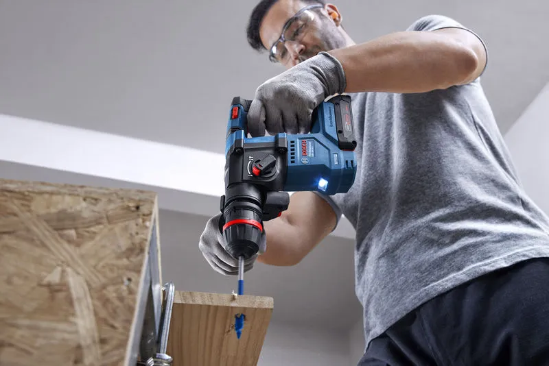 A person wearing safety equipment drills into wood with a cordless rotary hammer.