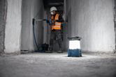 A worker wearing safety equipment drills a wall, cordless jobsite light on the floor.