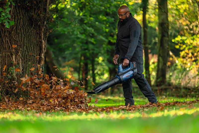 Person wearing safety equipment uses a leaf blower to clear fallen leaves near a tree.