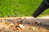 Power blower nozzle moves dry leaves across a gravel path outdoors.