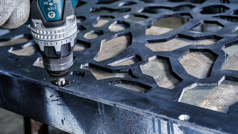 Person wearing safety equipment uses a cordless drill to make a hole in a metal sheet.