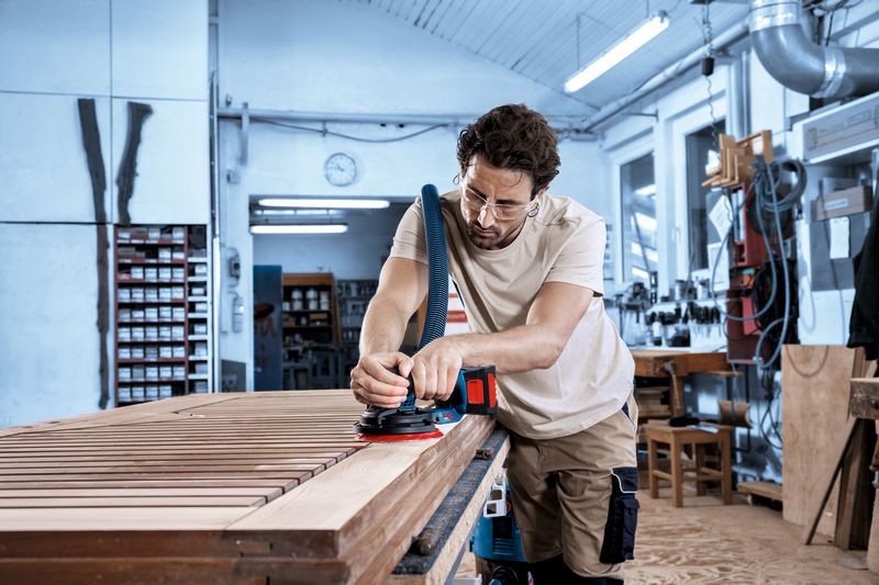 A person wearing safety equipment sands a wooden surface with a cordless random orbit sander.