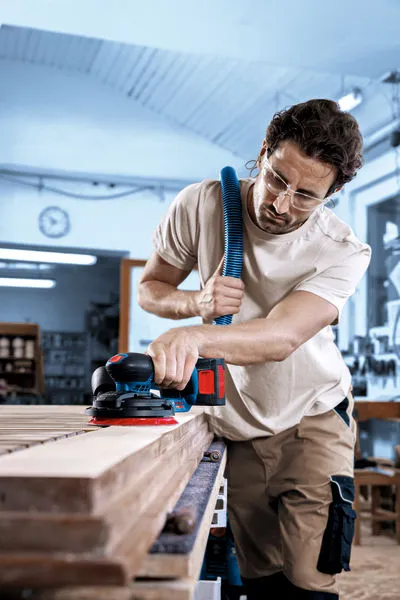 A person wearing safety equipment sands wooden boards with a cordless random orbit sander.