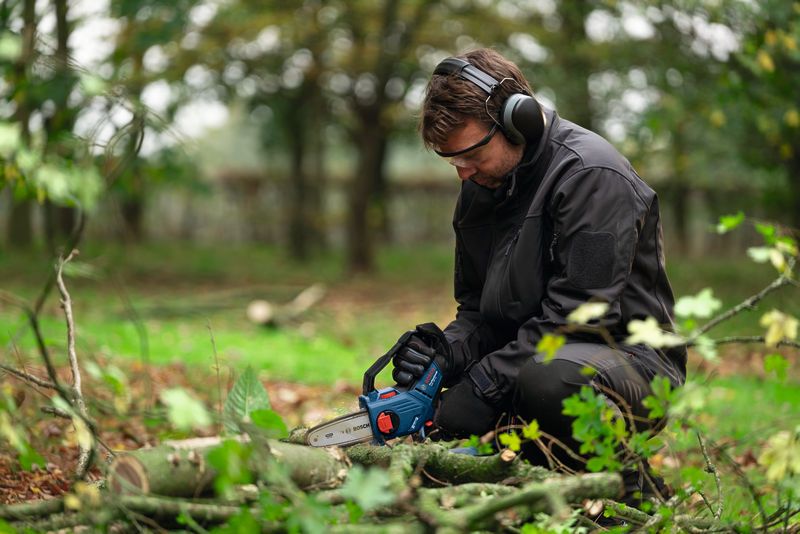 A person wearing safety equipment cuts tree branches with a chainsaw outdoors.