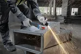 A person wearing safety equipment grinds a metal duct with an angle grinder, producing sparks.