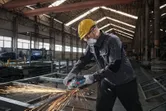 A person wearing safety equipment grinds metal with an angle grinder in a factory.