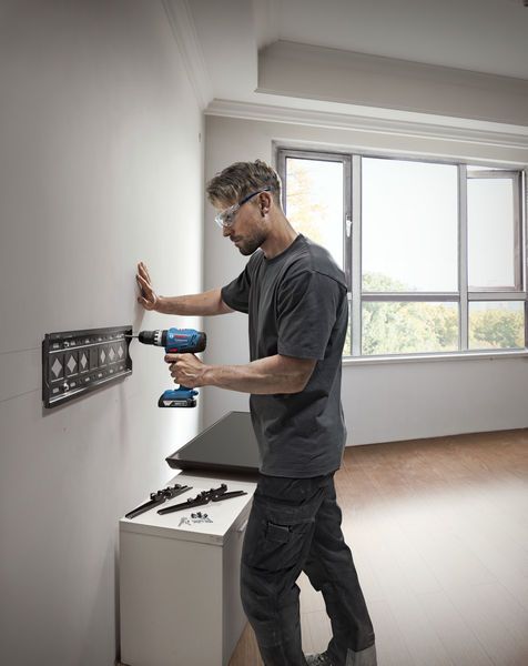 A person wearing safety equipment installs a metal bracket on a wall using a cordless drill.