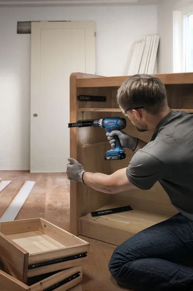 A person wearing safety equipment uses a cordless drill to assemble a wooden cabinet.