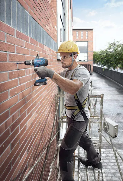 A person wearing safety equipment drills into a brick wall while standing on scaffolding.