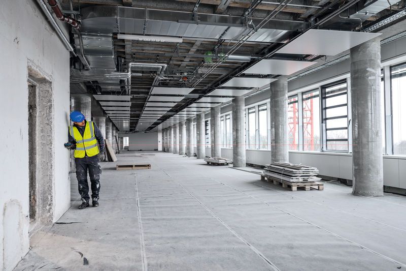 A person wearing safety equipment uses a laser measure to check wall distance in a large building.