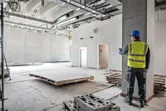 A person wearing safety equipment uses a laser measure in a large unfinished building.
