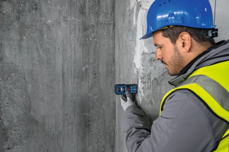 A person wearing safety equipment uses a laser measure against a concrete wall.
