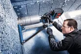 A person wearing safety equipment operates a mounted diamond drill on a concrete wall.