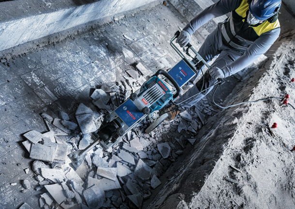 Person wearing safety equipment breaks concrete floor using a heavy-duty demolition tool.