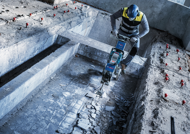 Person wearing safety equipment removes pool tiles using a heavy-duty demolition tool.