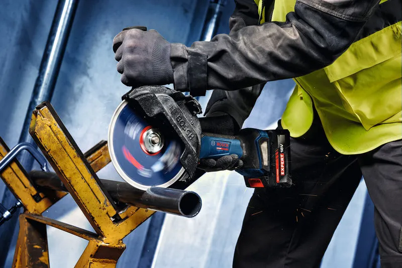 A person wearing safety equipment cuts a metal pipe using a cordless angle grinder.