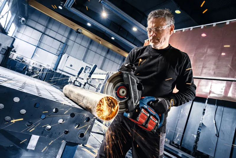 A person wearing safety equipment cuts a metal pipe with a cordless angle grinder.