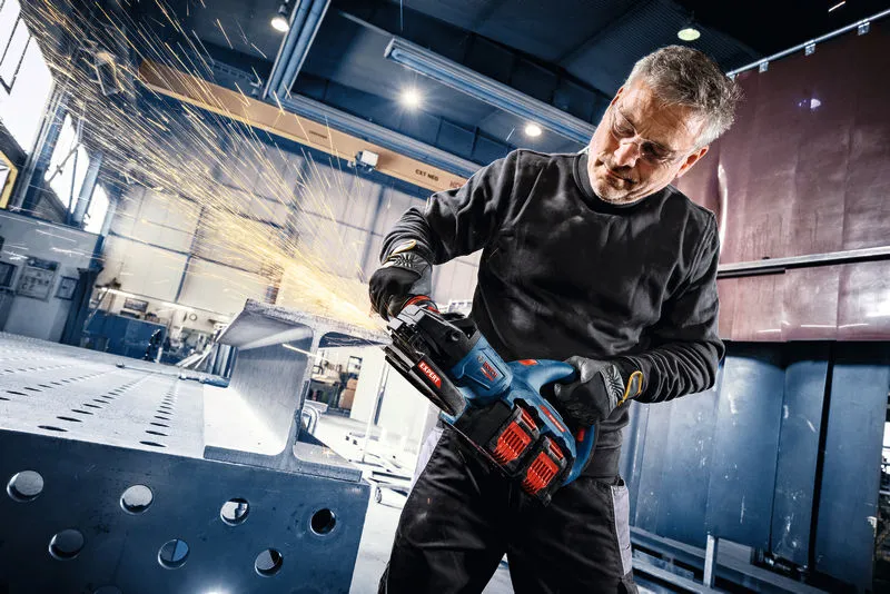 A person wearing safety equipment grinds a steel beam with a cordless angle grinder.