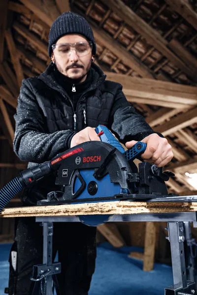 A person wearing safety equipment operates a cordless circular saw to cut wood.