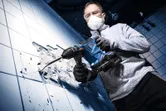 A person wearing safety equipment drills into a tiled wall using a cordless rotary hammer.