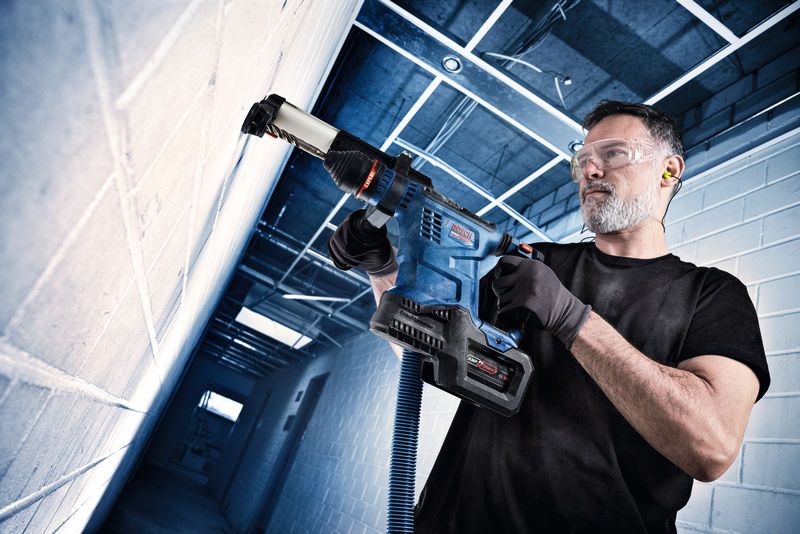 A person wearing safety equipment drills into a concrete wall using a cordless rotary hammer.