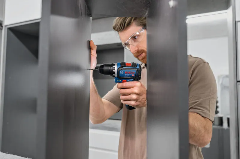 A person wearing safety equipment drills into a dark cabinet panel.
