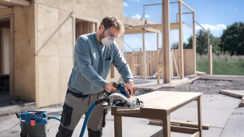 A person wearing safety equipment cuts wood with a cordless circular saw at a construction site.