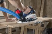 A person guides a cordless circular saw with dust extraction hose while cutting plywood.