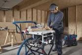 A person wearing safety equipment makes a wood cut using a table saw in a workshop.