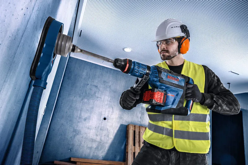A person wearing safety equipment drills into a concrete wall using a cordless rotary hammer.