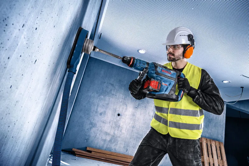 A person wearing safety equipment drills into a concrete wall using a cordless rotary hammer.