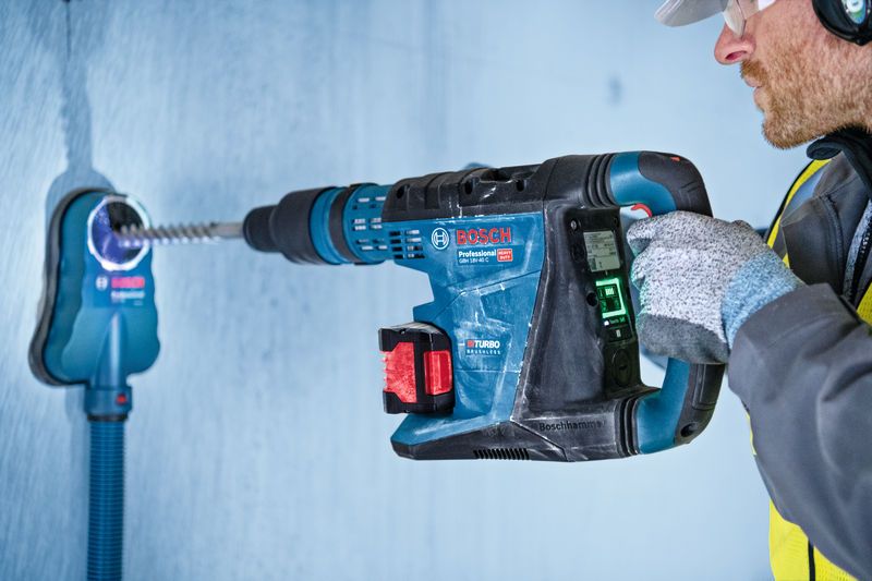 A person wearing safety equipment operates a rotary hammer drill with dust extraction.