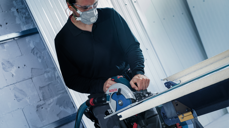 Person wearing safety equipment cuts a board with a circular saw on a workbench.