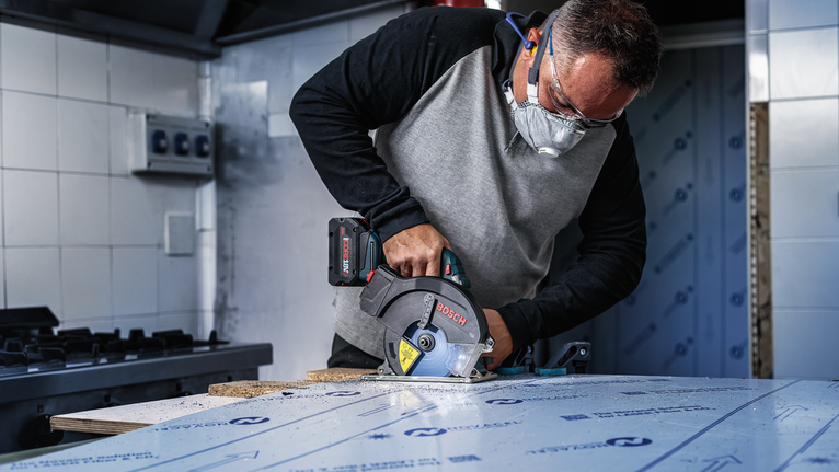 Person wearing safety equipment cuts laminate board with a cordless circular saw.