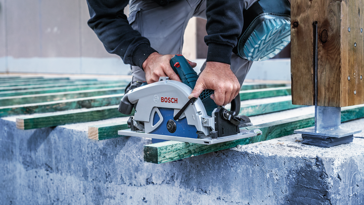 Person wearing safety equipment cuts a wooden beam with a circular saw on a construction site.
