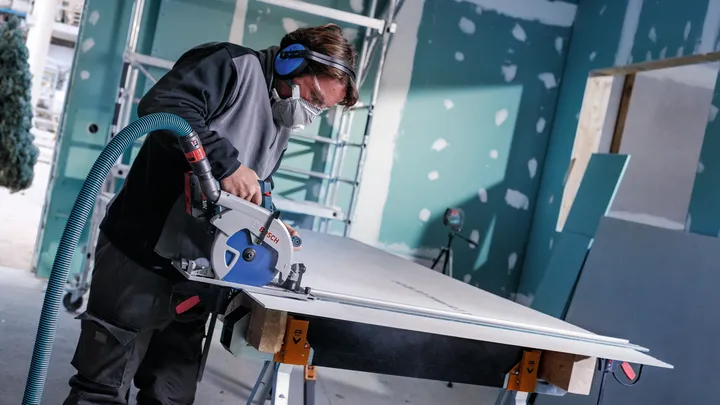 A person wearing safety equipment cuts drywall on a workbench with a power saw.