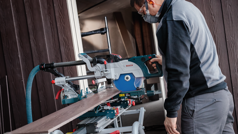 Person wearing safety equipment cuts a wooden board using a sliding miter saw.