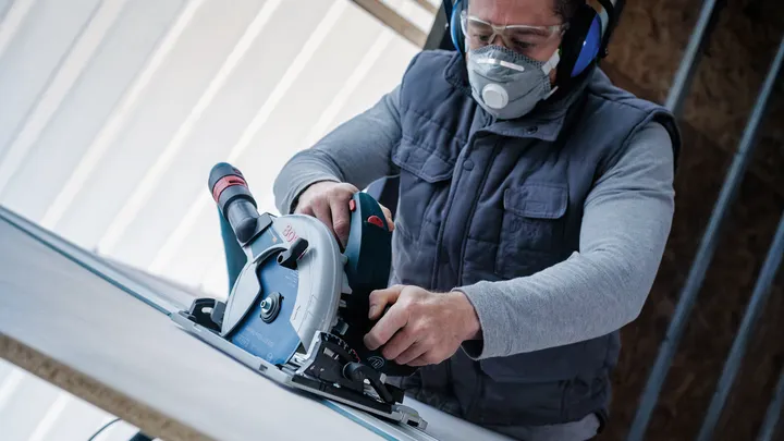 Person wearing safety equipment uses a circular saw to cut a board.