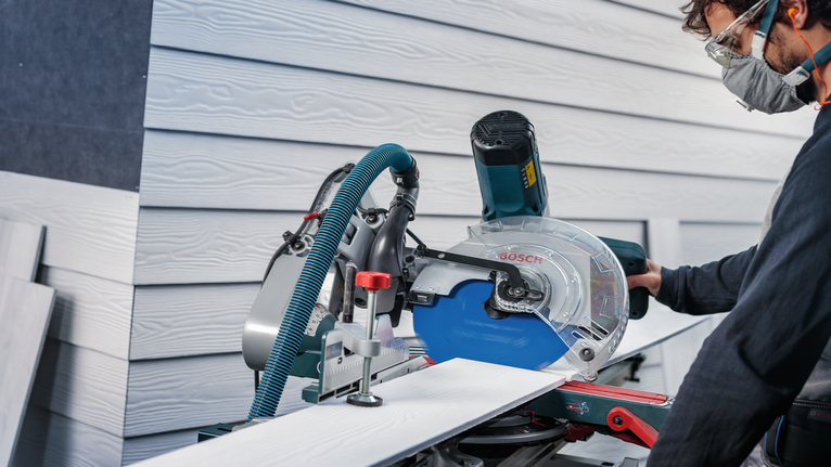 Person wearing safety equipment operates a miter saw to cut siding boards.