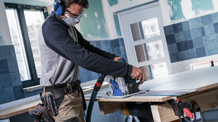 Person wearing safety equipment cuts a board with a power saw on a workbench.