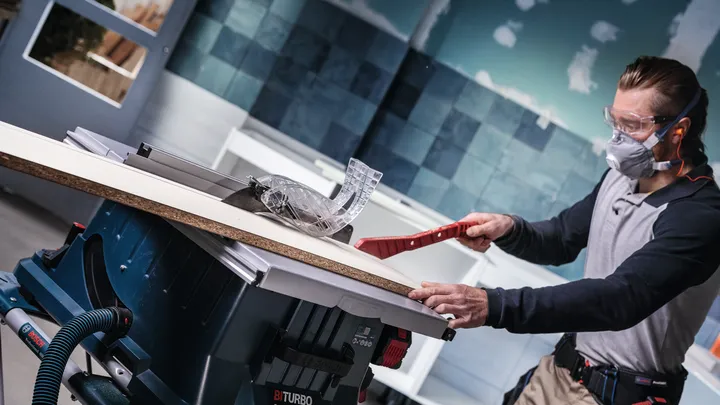 Person wearing safety equipment guides wood through a table saw in a workshop.