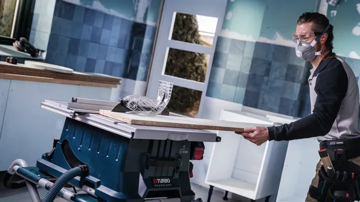 Person wearing safety equipment cuts a wooden board on a table saw in a workshop.