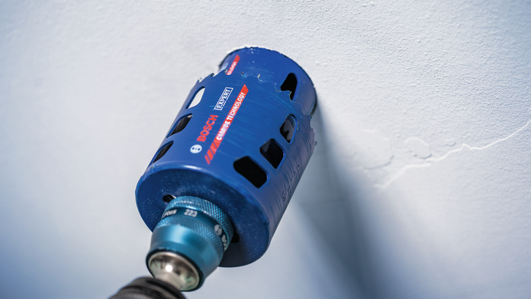 Power tool with a blue hole saw attachment drills into a white wall.