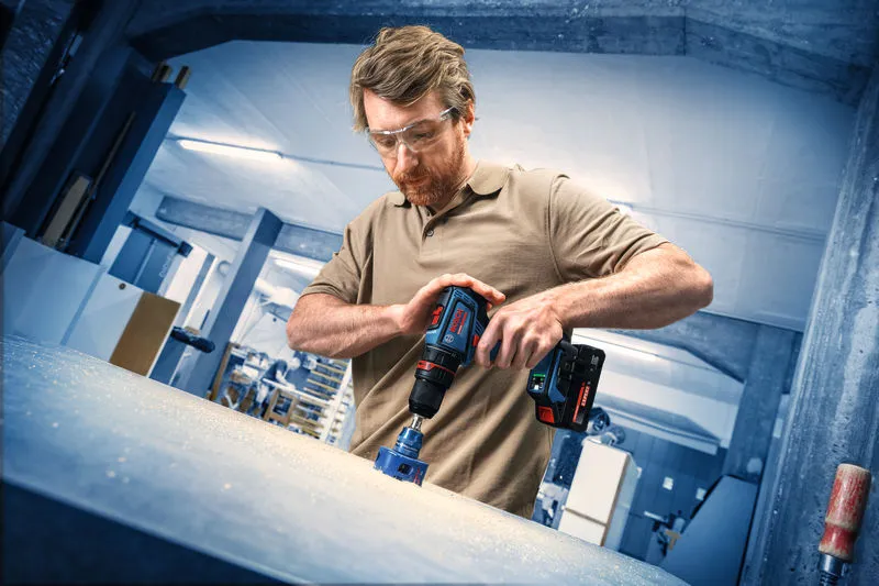 A person wearing safety equipment drills into a work surface with a cordless drill.