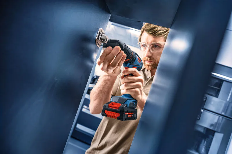 A person wearing safety equipment uses a cordless drill driver in a metal structure.