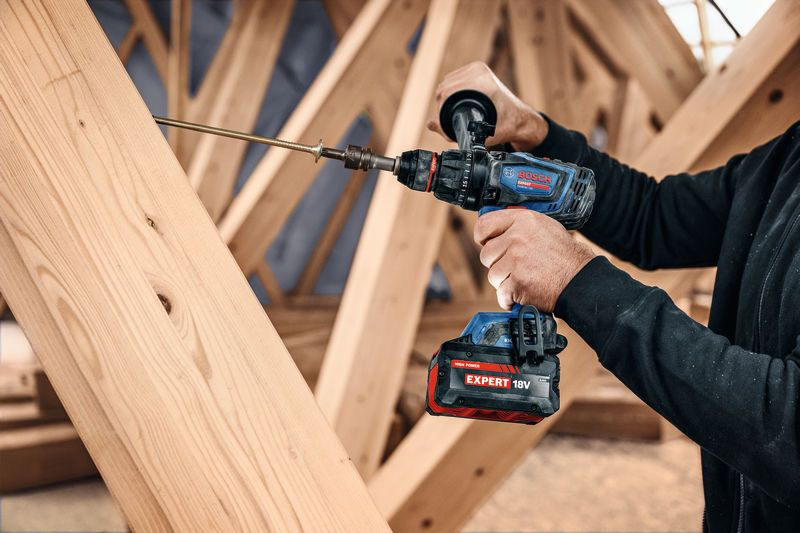 A person drives a long screw into wooden beams using a cordless combi drill.