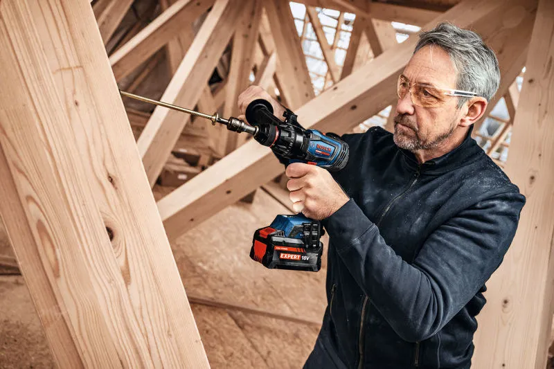 A person wearing safety equipment uses a cordless drill to drive a screw into wooden beams.